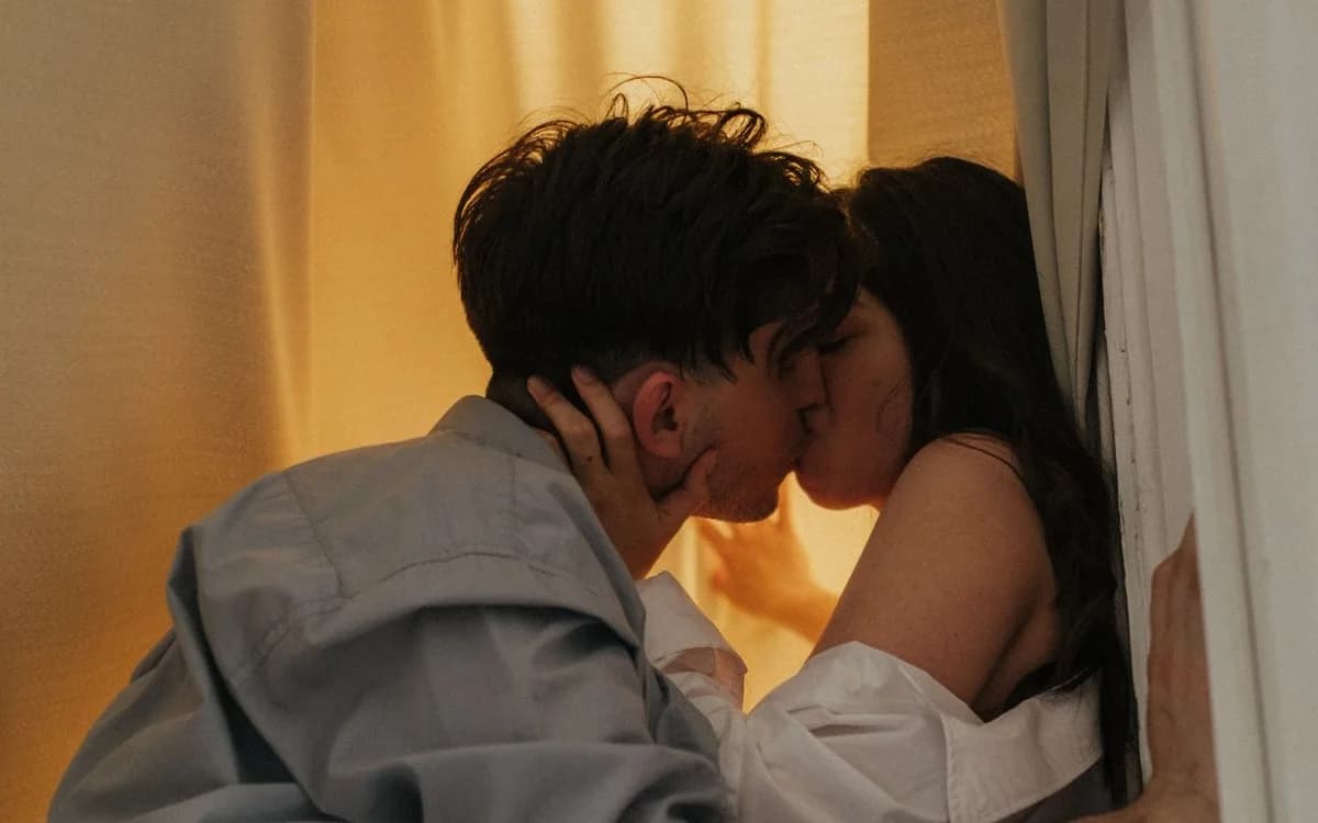 An adult couple sharing a close kiss beside curtains in warm evening light.